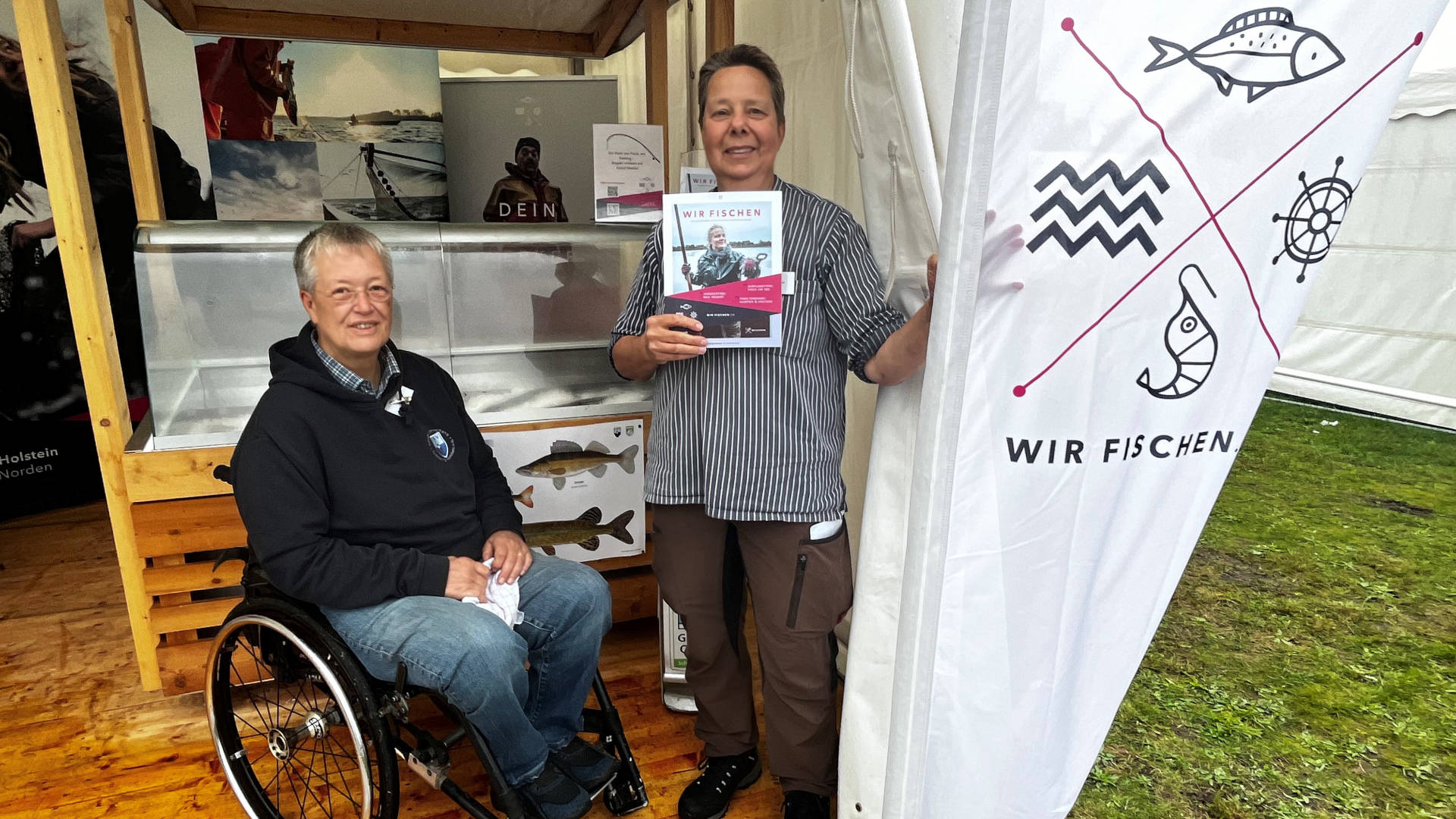 Eine Frau im Rollstuhl und eine stehende Frau mit Zeitschrift in der Hand