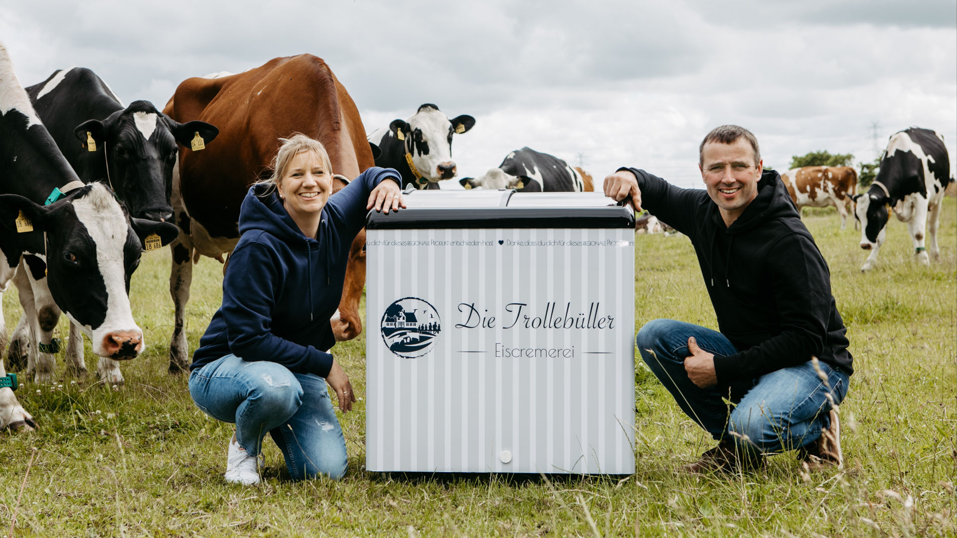 Milchkühe auf der Weide, im Vordergrund ein Mann und eine Frau mit einer mobilen Eistruhe