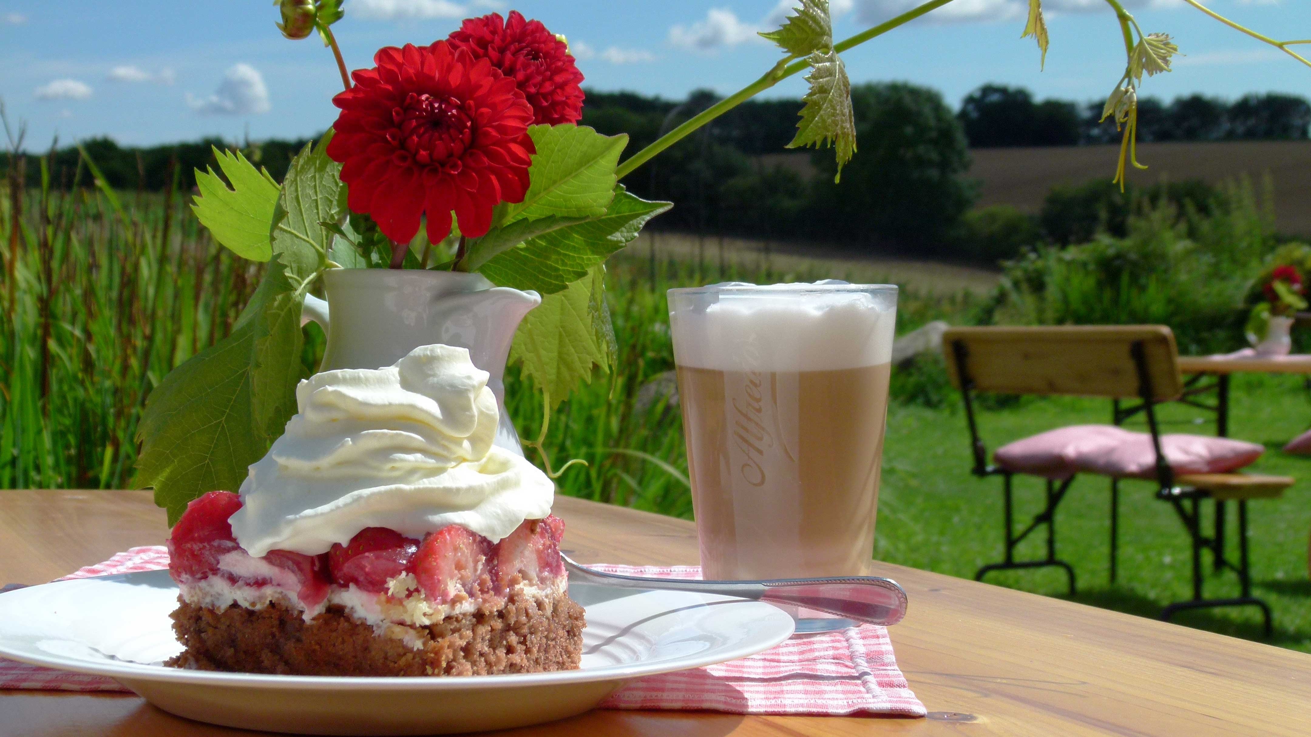 Ein Stück Erdbeerkuchen und ein Milchkaffe stehen auf einem Tisch, gemeinsam mit einer Blumenvase mit roten Blumen. Im Hintergrund sind die Weinberge zu sehen