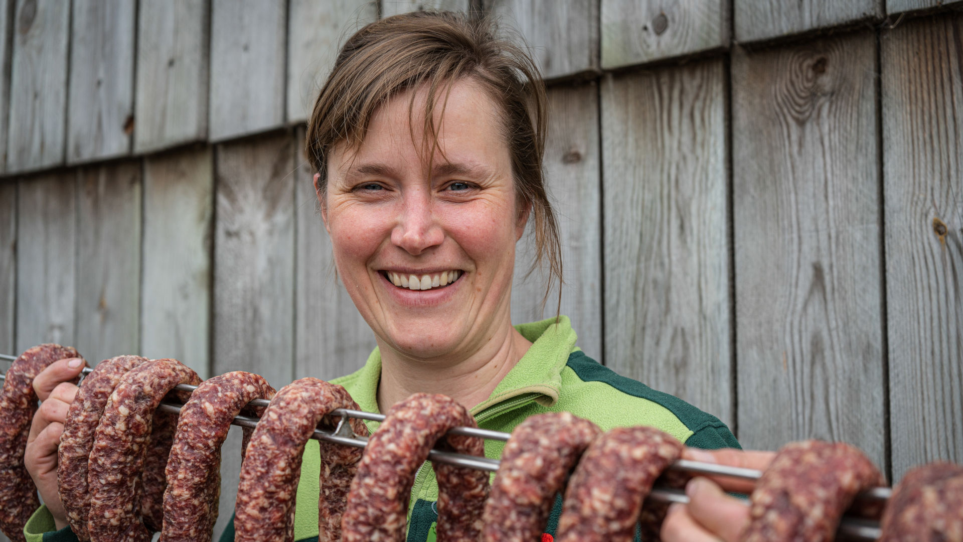 Frau lächelt in die Kamera, hält eine Stande mit luftgetrockneter Salami in die Höhe