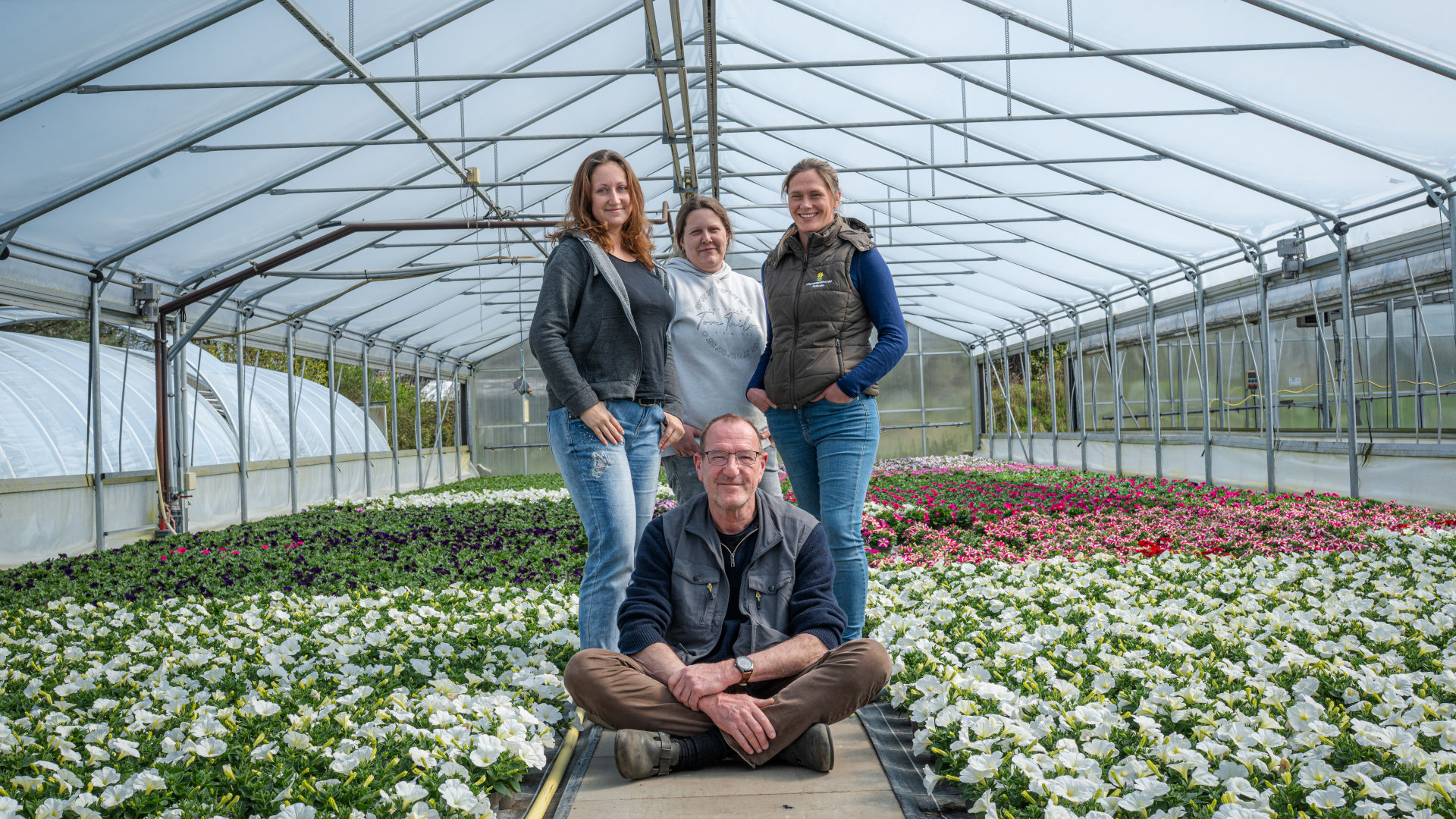 ein Mann sitzt auf dem Boden eines Gewächshauses, drei Frauen stehen hinter ihm, überall stehen Blumen