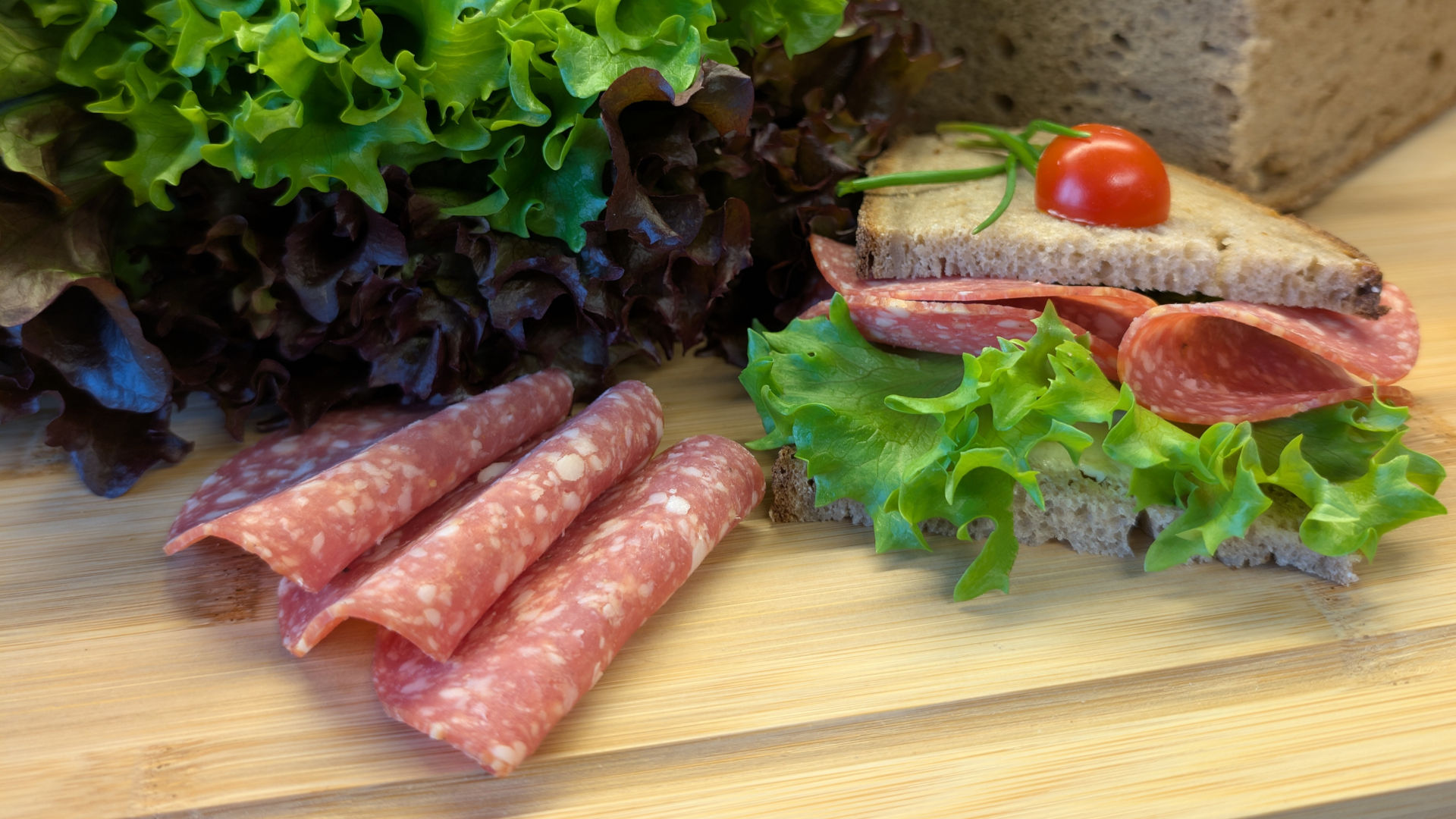 Salami-Stulle auf einem Holzbrett, dekoriert mit Tomate, im Hintergrund ein Salatkopf, daneben dekorierte Salamischeiben