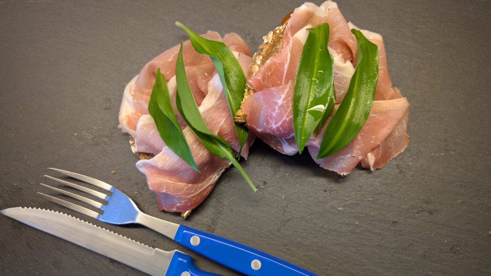zwei Stullen mit Schinken und Bärlauch auf einer Schieferplatte, daneben Besteck