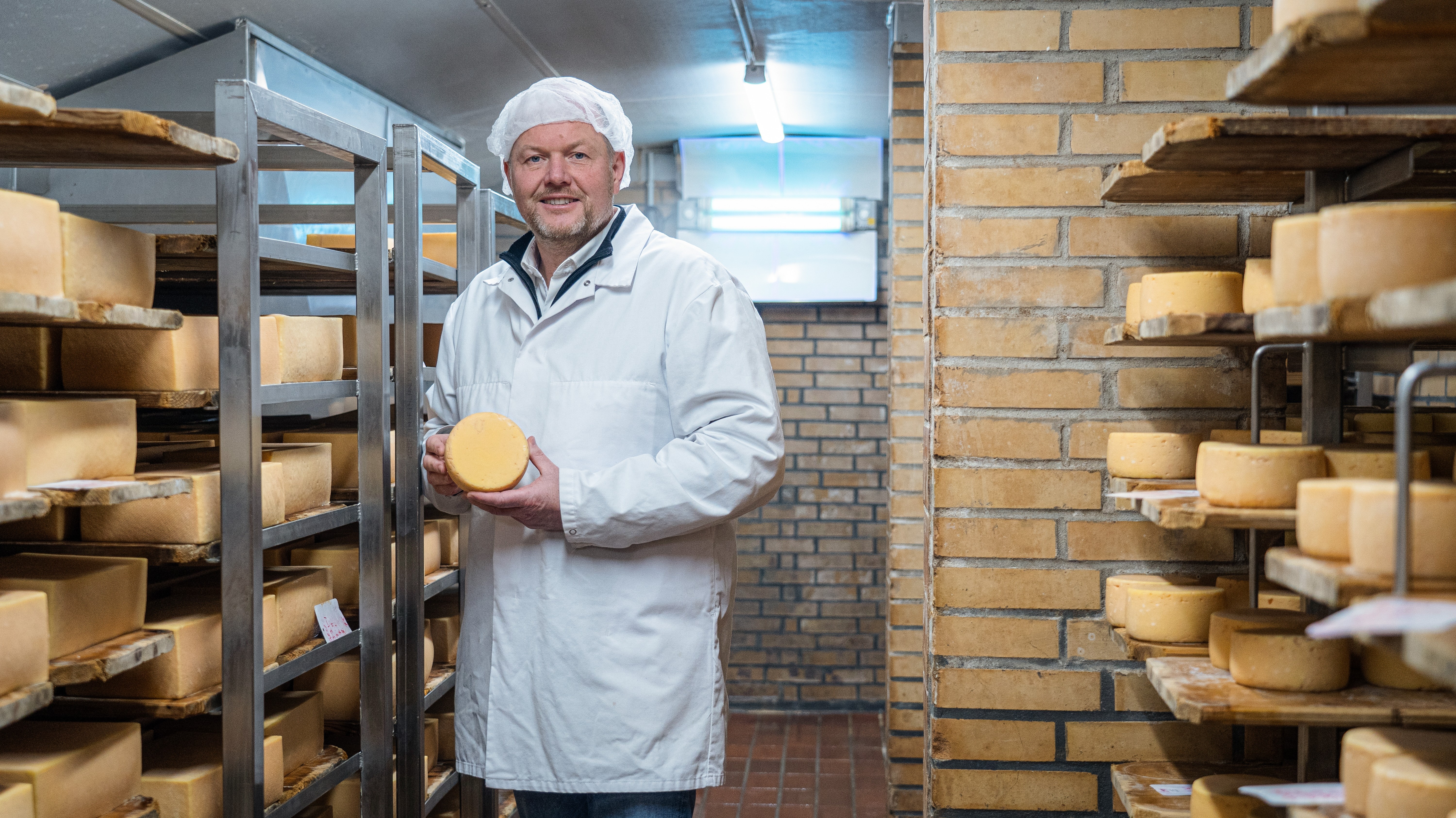 Mann steht in Käsereifekammer, hält einen Käse in der Hand